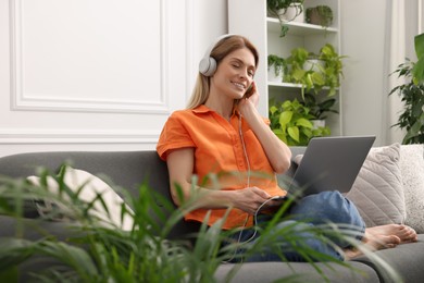 Photo of Woman in headphones using laptop on sofa near beautiful potted houseplants at home