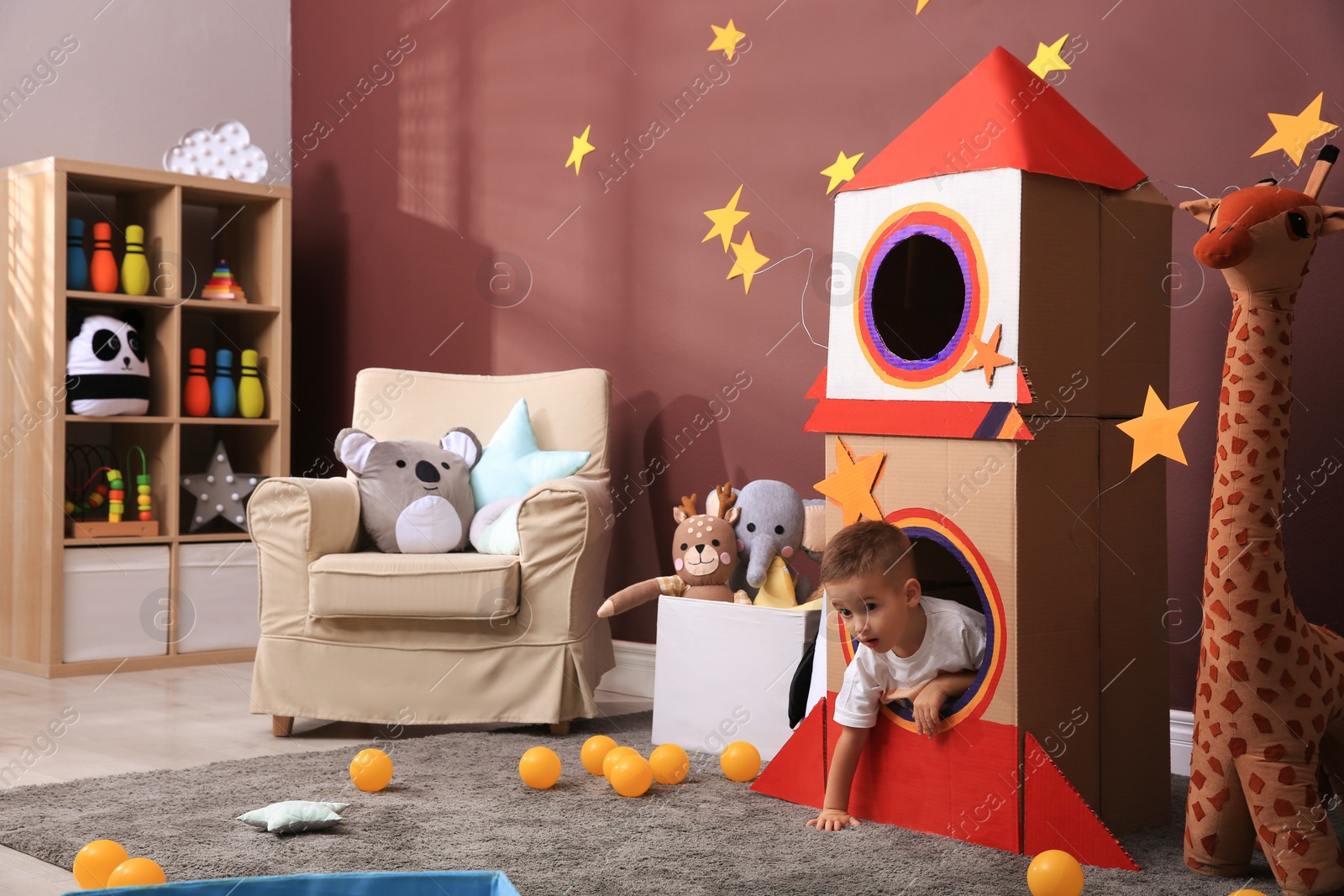 Photo of Cute little boy playing with cardboard rocket and toys at home. Child's room interior