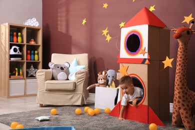 Photo of Cute little boy playing with cardboard rocket and toys at home. Child's room interior