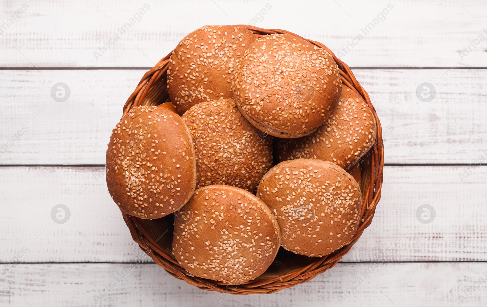 Photo of Wicker basket of fresh buns with sesame seeds on white wooden table, top view