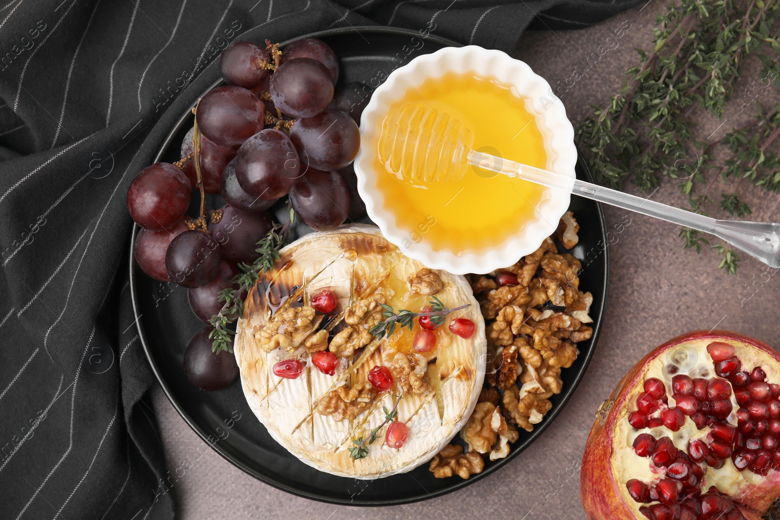 Photo of Plate with tasty baked camembert, honey, grapes, walnuts and pomegranate seeds on brown textured table, top view