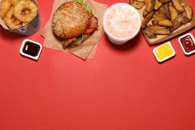 Tasty burger, potato wedges, fried onion rings, different sauces and refreshing drink on red background, flat lay with space for text. Fast food