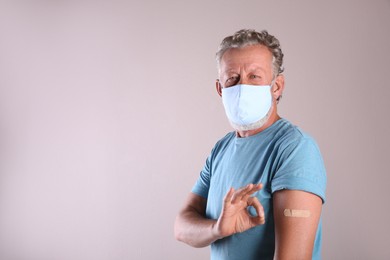 Cheerful senior man in protective mask showing arm with bandage after vaccination on beige background. Space for text