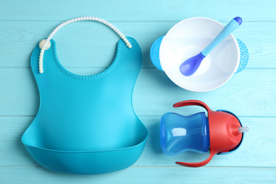 Set of plastic dishware and silicone bib on light blue wooden table, flat lay. Serving baby food