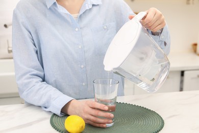 Photo of Woman pouring water from filter jug into glass at white marble table in kitchen, closeup