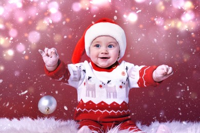 Cute baby in Santa hat with Christmas ball on fluffy carpet against red background. Magical festive atmosphere