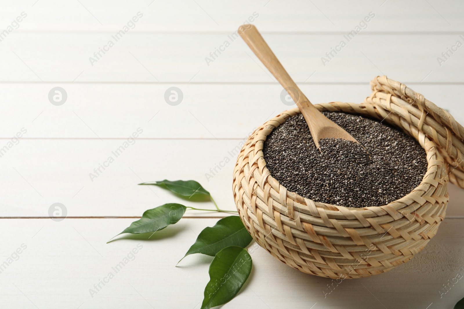 Photo of Wicker box with chia seeds and spoon on white wooden table. Space for text