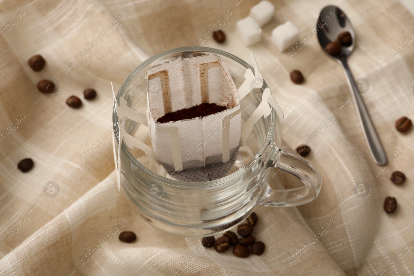 Photo of Glass cup with drip coffee bag and beans on beige fabric, closeup