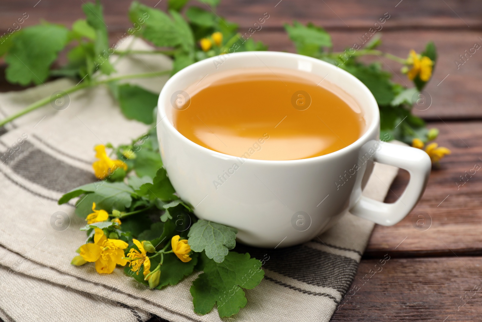 Photo of Cup of aromatic celandine tea and flowers on wooden table, closeup