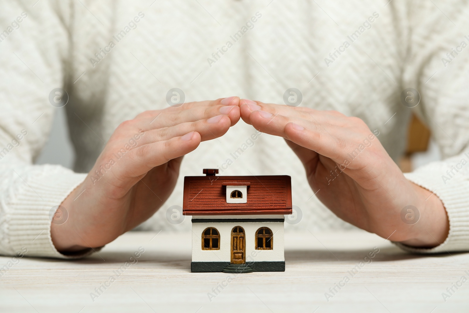 Photo of Home security concept. Man covering house model at white wooden table, closeup
