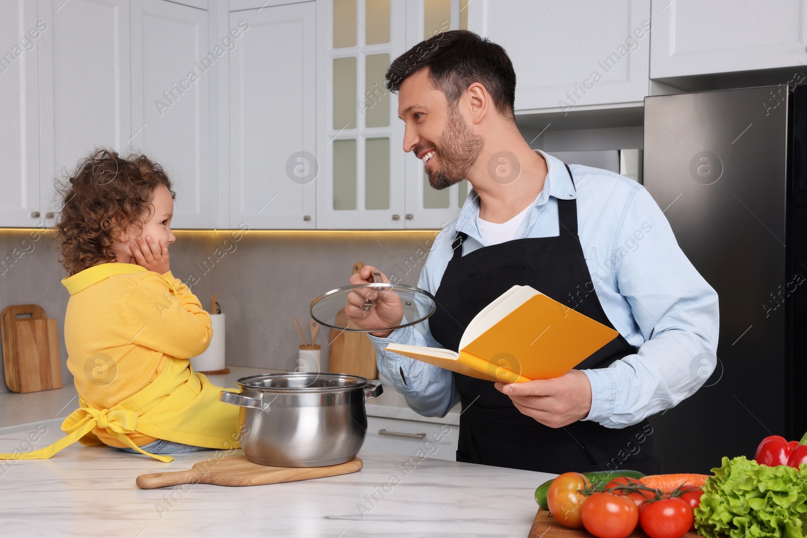 Photo of Cute little girl and her father with recipe book cooking in kitchen