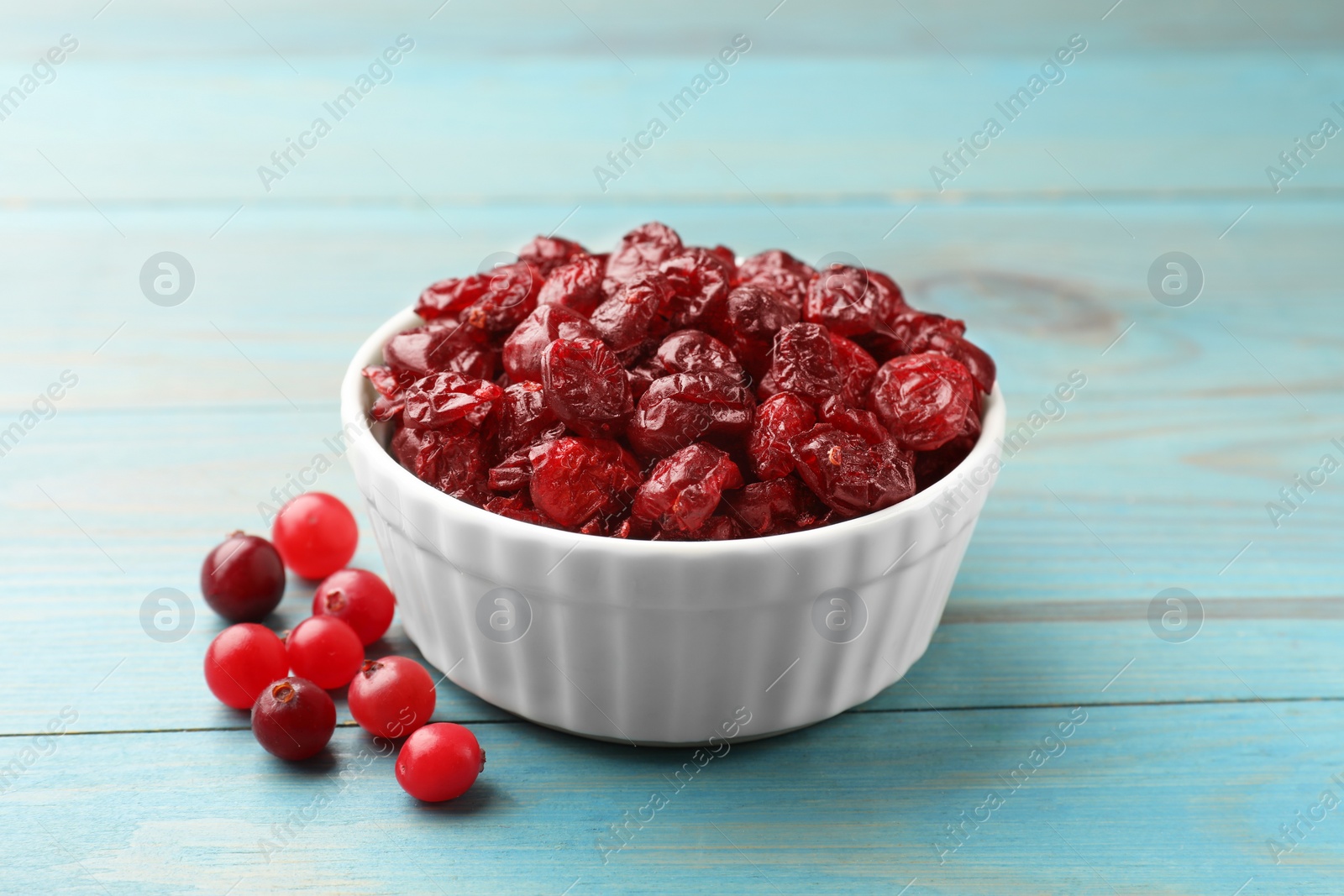 Photo of Tasty dried cranberries in bowl and fresh ones on light blue wooden table, closeup