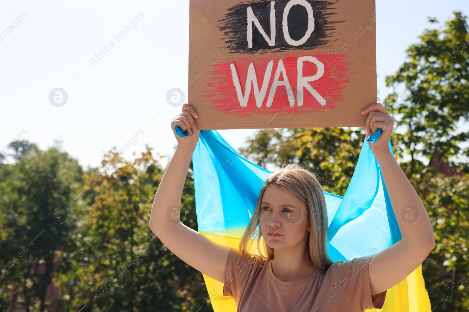 Photo of Sad woman holding poster with words No War and Ukrainian flag outdoors