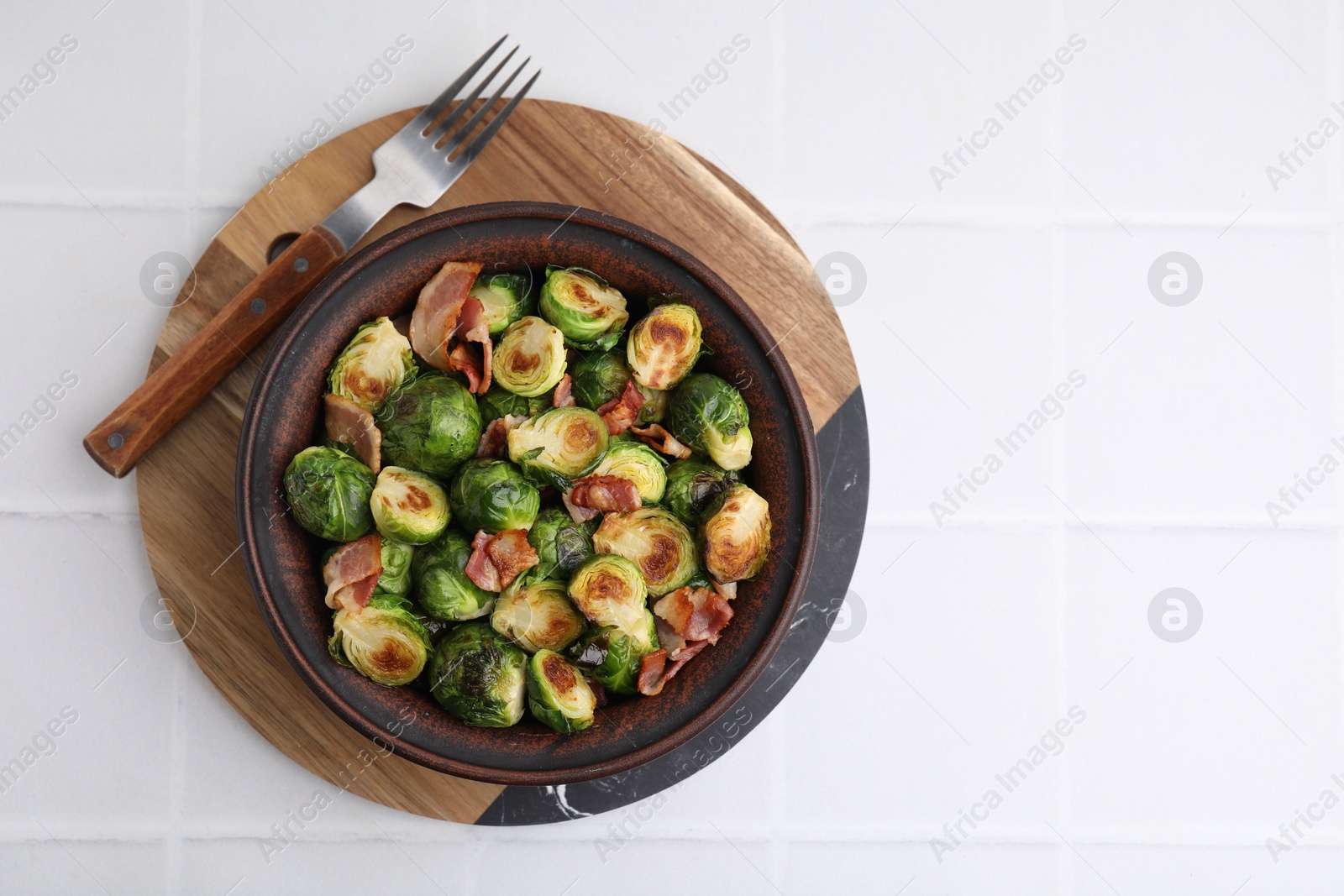 Photo of Delicious roasted Brussels sprouts and bacon in bowl on white tiled table, top view. Space for text