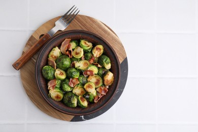 Photo of Delicious roasted Brussels sprouts and bacon in bowl on white tiled table, top view. Space for text