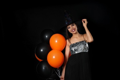 Beautiful woman wearing witch costume with balloons for Halloween party on black background, space for text