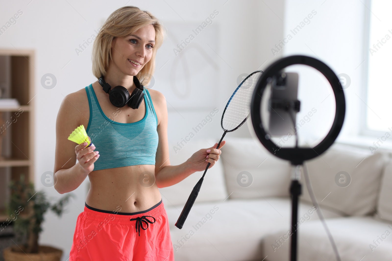 Photo of Smiling sports blogger holding badminton racket and shuttlecock while streaming online fitness lesson with smartphone at home