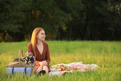Beautiful young woman with picnic basket sitting on blanket in park