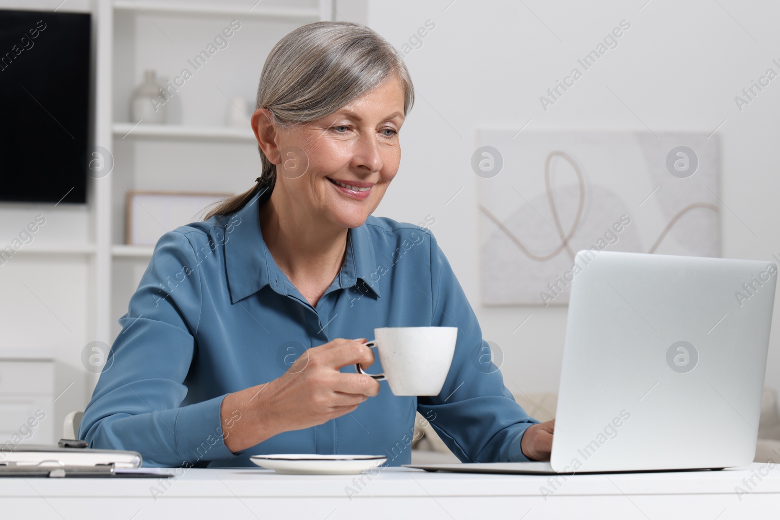 Photo of Beautiful senior woman with cup of drink using laptop at white table indoors