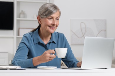 Photo of Beautiful senior woman with cup of drink using laptop at white table indoors