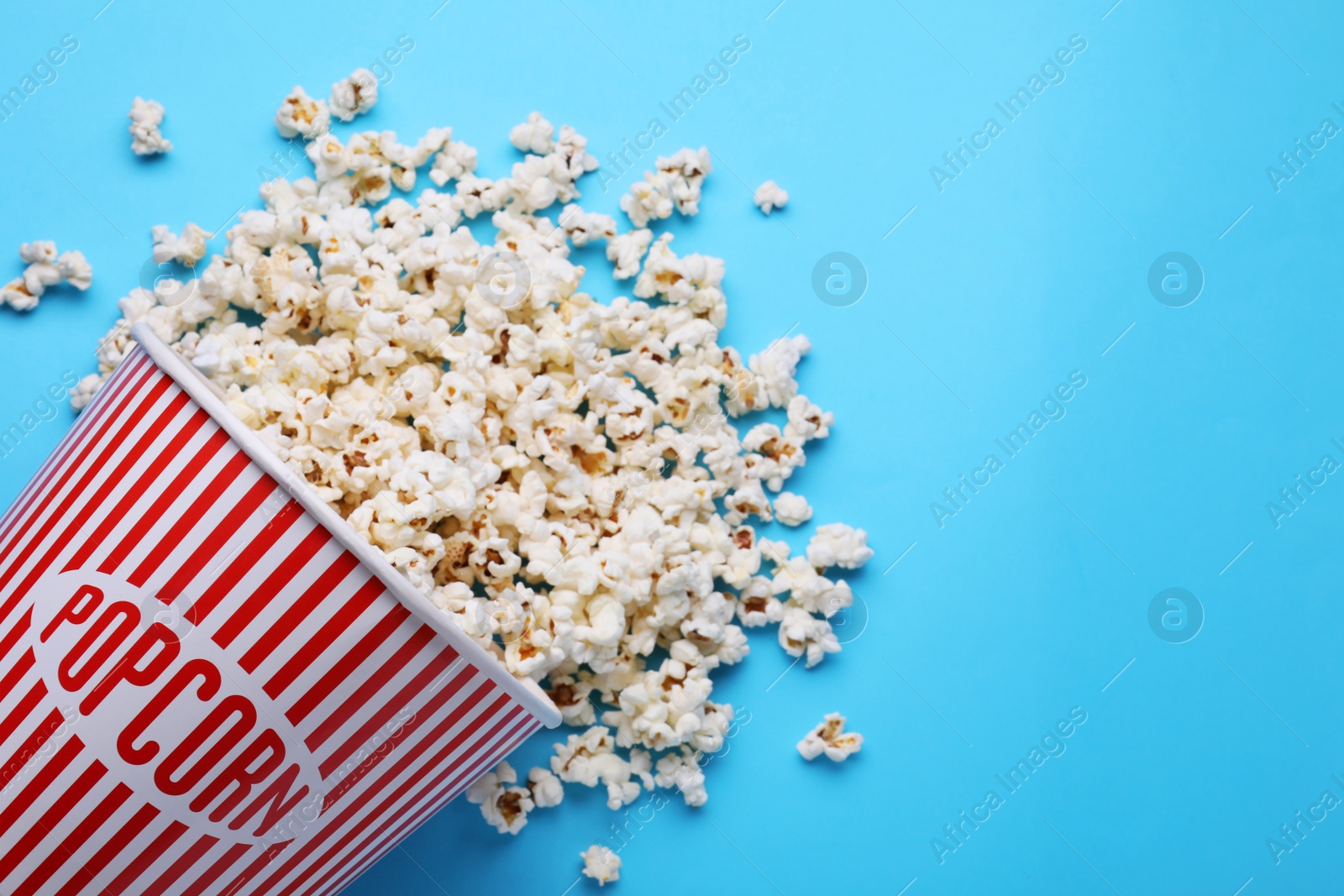 Photo of Overturned paper bucket with delicious popcorn on light blue background, flat lay. Space for text