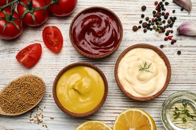 Photo of Bowls with mustard, ketchup, mayonnaise and ingredients on wooden table, flat lay