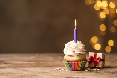 Photo of Birthday cupcake with candle and gift box on wooden table against blurred lights. Space for text