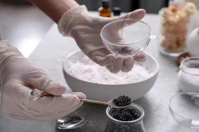 Photo of Woman in gloves filling bath bomb mold with dried flower buds at grey table, closeup