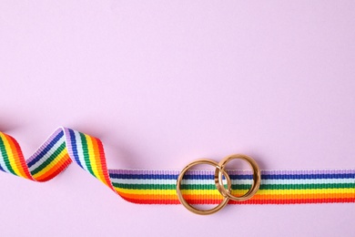 Top view of wedding rings and rainbow ribbon on color background, space for text. Gay symbol