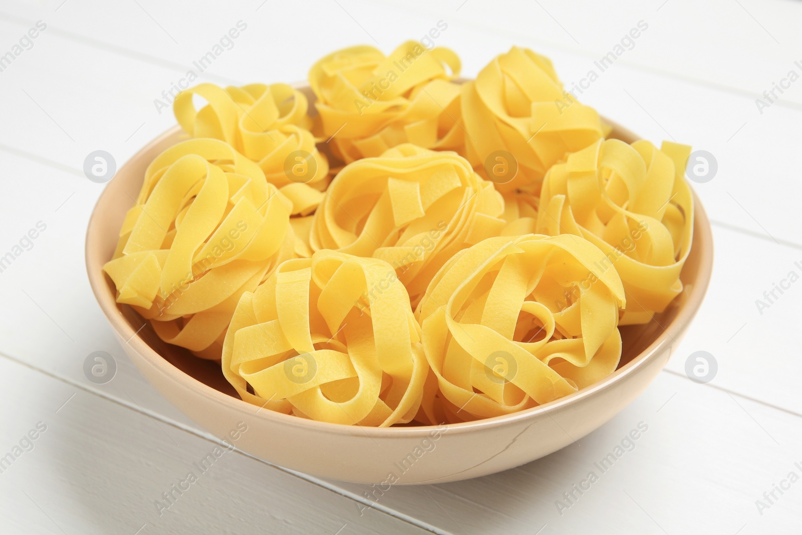 Photo of Raw tagliatelle pasta in bowl on white wooden table, closeup