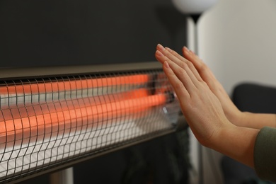 Woman warming hands near electric infrared heater indoors, closeup