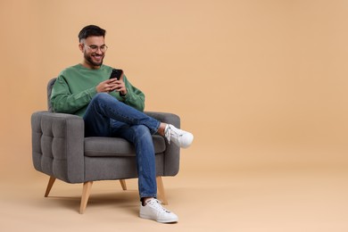 Photo of Happy young man using smartphone in armchair on beige background, space for text