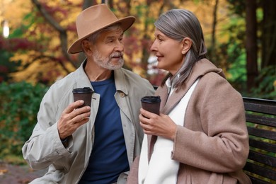 Affectionate senior couple with cups of coffee spending time together on bench in autumn park