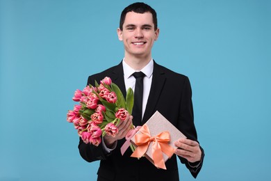 Happy young man with beautiful bouquet and present on light blue background