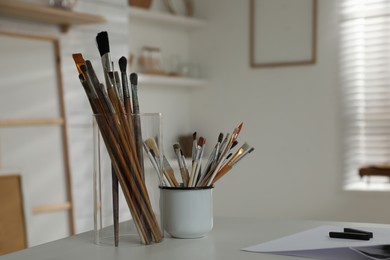 Photo of Holders with different paintbrushes on white table in studio, space for text. Artist's workplace