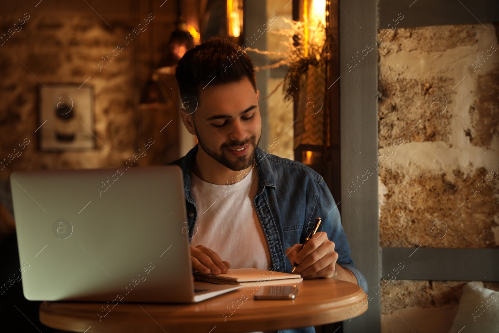 Photo of Young blogger writing in notebook at table indoors