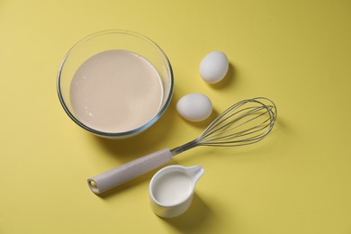 Photo of Metal whisk, dough in bowl, eggs and milk on pale yellow background, above view