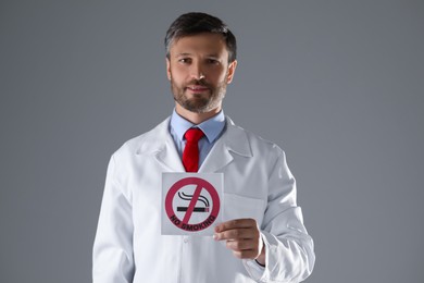 Photo of Doctor holding card with no smoking sign on gray background, selective focus