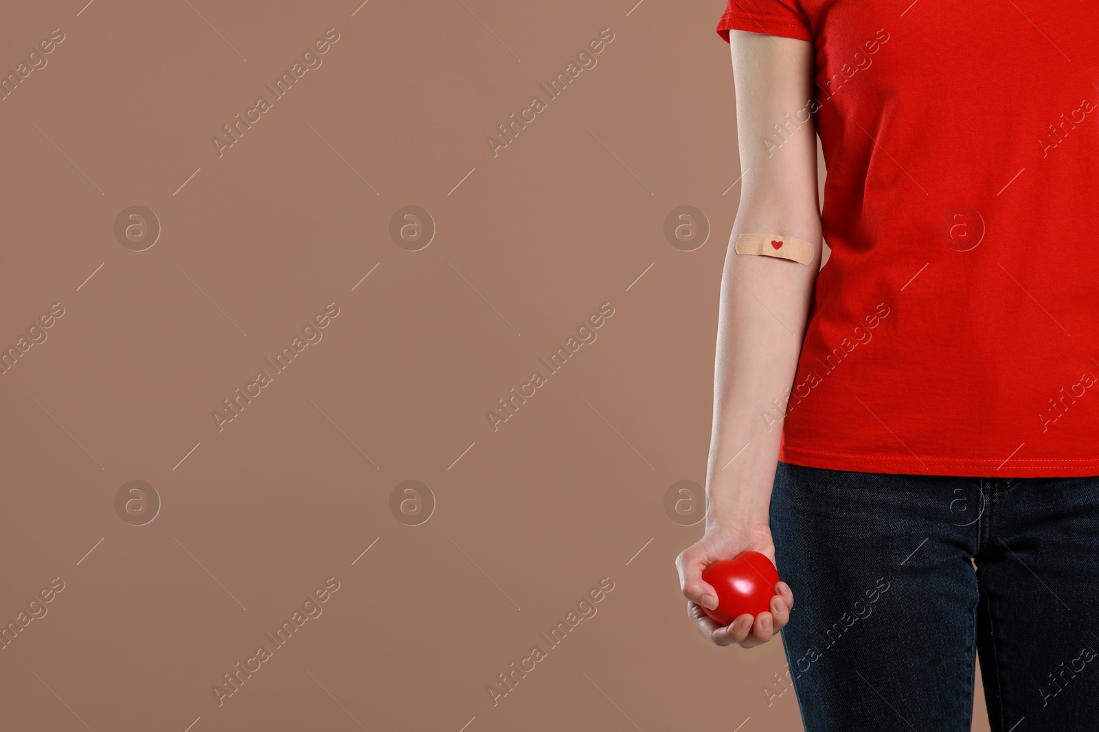 Photo of Blood donation concept. Woman with adhesive plaster on arm holding red heart against brown background, closeup. Space for text