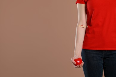 Blood donation concept. Woman with adhesive plaster on arm holding red heart against brown background, closeup. Space for text