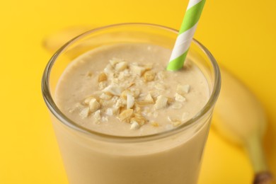 Glass of tasty banana smoothie with straw and nuts on yellow background, closeup