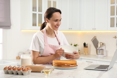 Photo of Woman making cake while watching online cooking course via laptop in kitchen