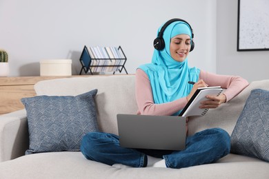 Photo of Muslim woman in headphones writing notes near laptop at couch in room