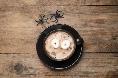 Photo of Cup of coffee decorated with marshmallow eyes on wooden table, flat lay. Halloween celebration