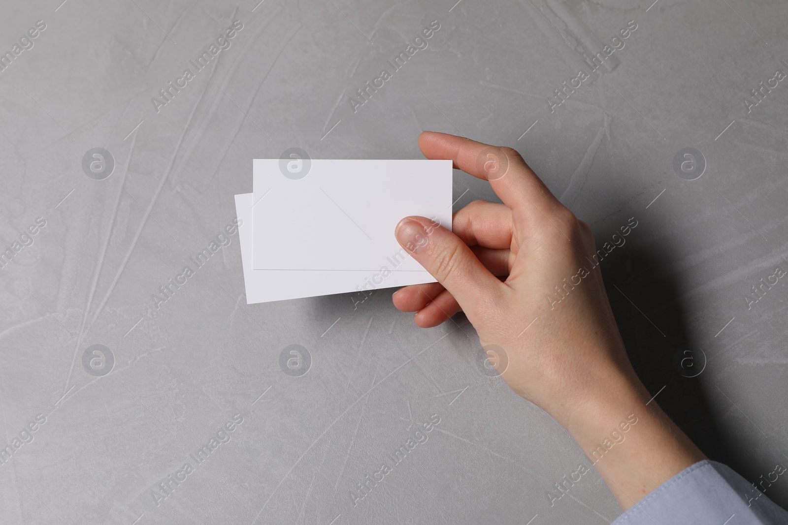 Photo of Woman holding blank cards at light grey table, top view. Mockup for design