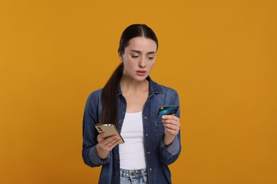 Confused woman with credit card and smartphone on orange background. Debt problem