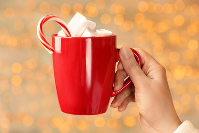 Woman holding cup of cacao with marshmallows and candy cane against blurred lights, closeup. Bokeh effect