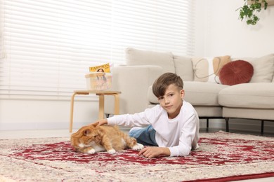 Photo of Little boy petting cute ginger cat on carpet at home