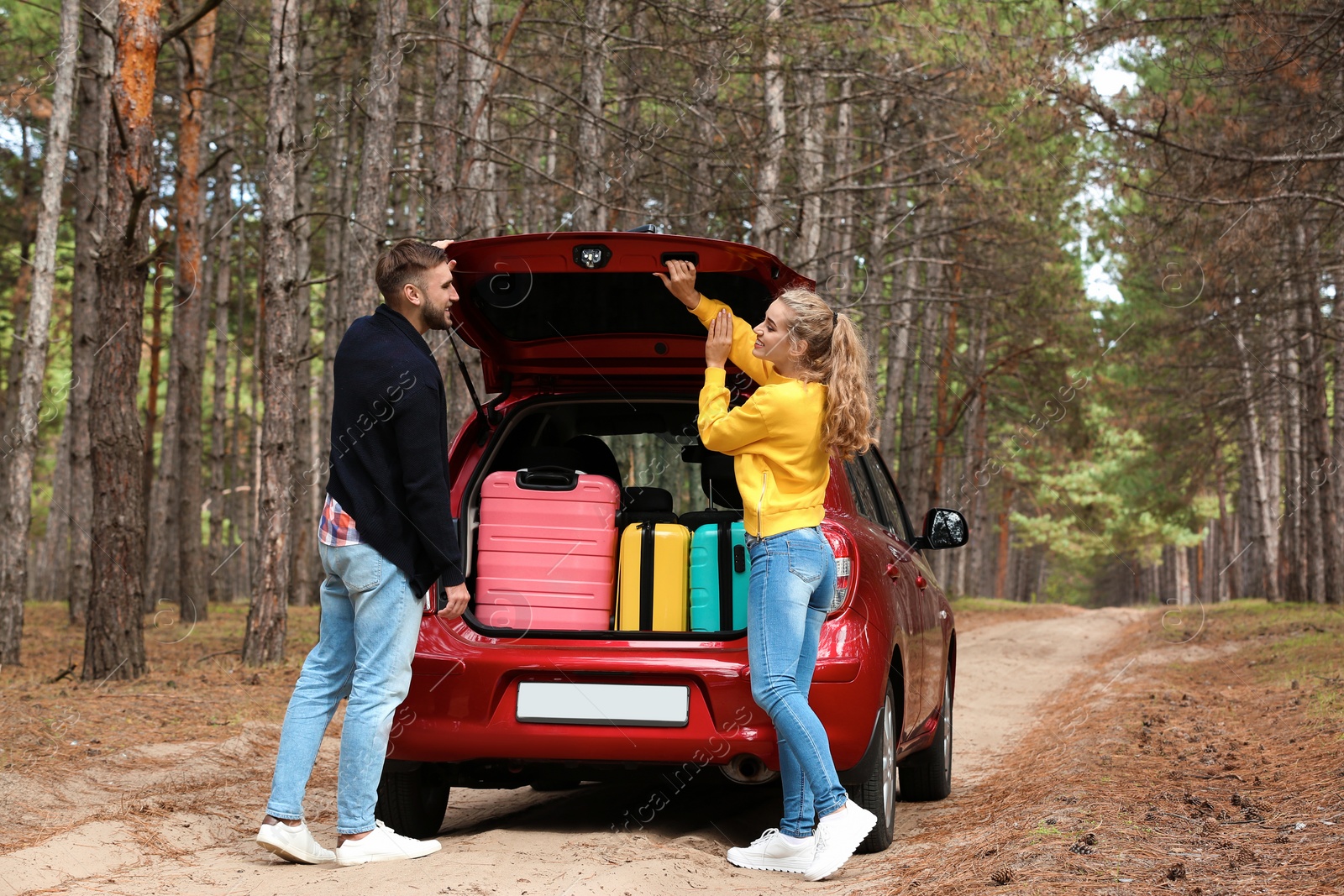 Photo of Young couple standing near car trunk loaded with suitcases on forest road