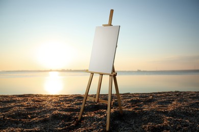 Image of Wooden easel with blank canvas on sandy beach near sea at sunrise 